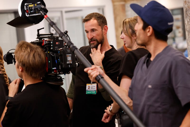 A film crew records a scene with actors wearing "Doctor" badges. One actor stands thoughtfully, touching his chin, while crew members operate a camera and boom mic on set.