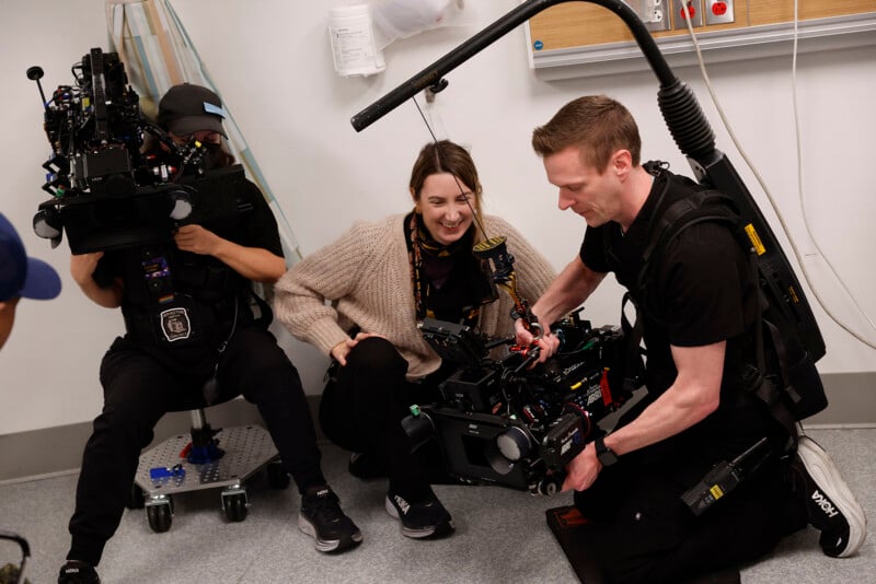 Two camera operators with professional gear crouch on a hospital set, smiling and adjusting equipment, while a woman between them laughs and looks at one of the cameras.