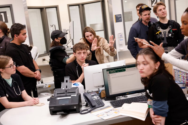 A film crew and actors gather around a hospital reception desk set, discussing a scene. Monitors, paperwork, and medical props are visible, with people focused on planning or listening intently.
