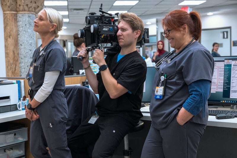 Three people in medical scrubs stand in a hospital setting; one holds a professional movie camera. Two women smile, while the man with the camera appears focused. Computer monitors and medical equipment are visible.
