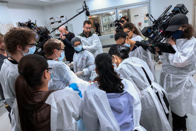 A group of people in medical gowns and masks gather around a patient in a hospital setting, while several camera operators film the scene. The atmosphere appears focused and collaborative.