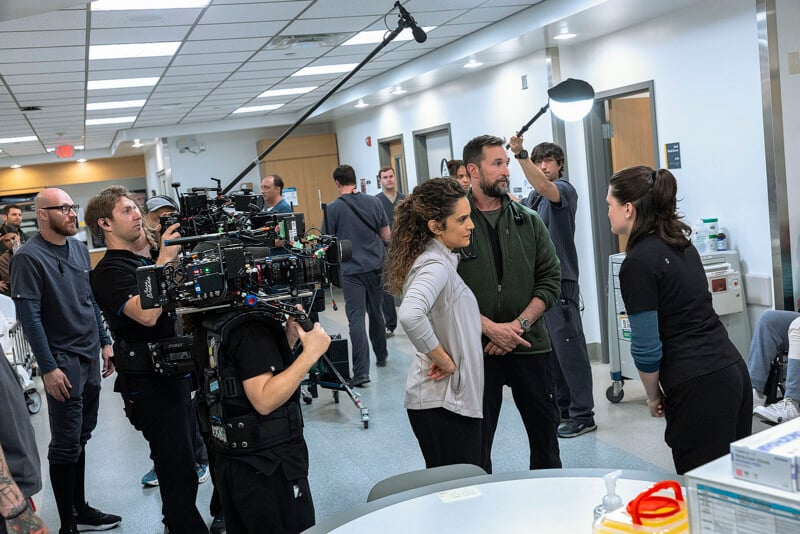 Film crew with cameras and boom mics shoot a scene in a busy hospital hallway, with actors in medical attire engaged in conversation while crew members watch and equipment fills the space.
