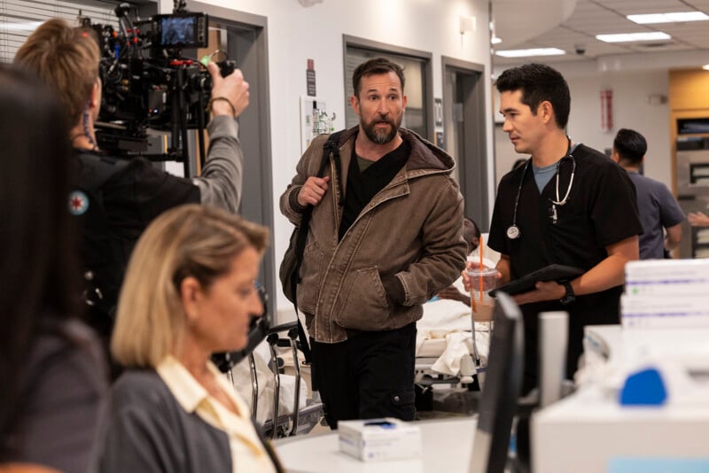 A film crew records a scene in a hospital setting, with one man in a brown jacket talking to a man in black scrubs with a stethoscope. Other people work in the background, and medical equipment is visible.