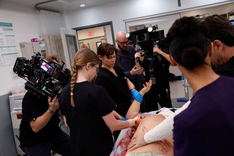 A film crew records actors and medical staff around a hospital bed during the filming of a medical scene, with cameras and crew members focused on the staged procedure.