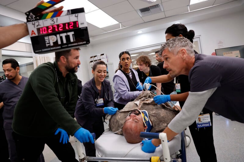 A film crew and actors on a hospital set surround an actor on a stretcher, preparing for a medical scene. A clapperboard is visible, and the team appears focused and engaged in their roles.