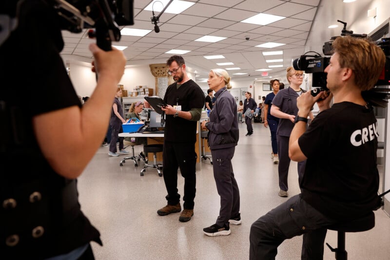 A film crew captures a scene in a busy hospital set, with actors in medical scrubs and crew members operating cameras and equipment in a well-lit room.