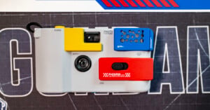 A close-up of a retro-style camera with yellow, blue, and red geometric elements on a gray body, placed on a dark surface with white and blue text and graphic designs.