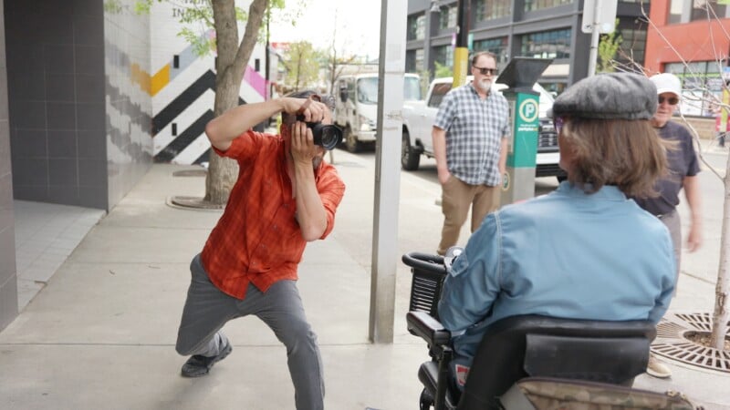 A photographer in an orange shirt crouches to take a photo of a person in a blue jacket in a mobility scooter. A man in a plaid shirt stands nearby.
