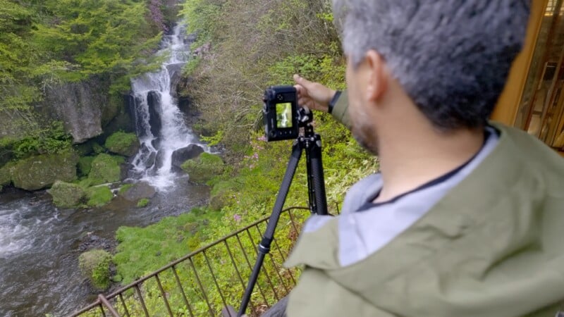 Un hombre de pelo gris y chaqueta verde se encuentra junto a una barandilla de metal rodeado de exuberante vegetación, fotografiando una cascada del bosque con una cámara montada en un trípode.