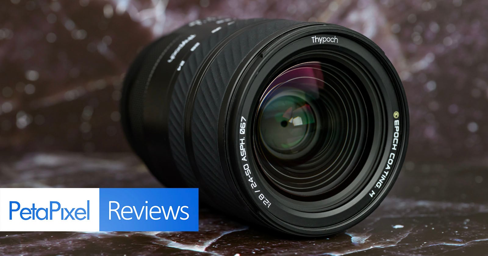 A close-up of a camera lens with visible reflections, lying on a dark marbled surface. The image includes a "PetaPixel Reviews" banner in the lower left corner.