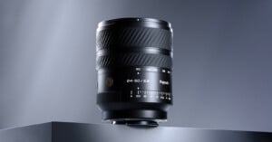 A black camera lens marked "24-50/2.8" and "TTArtisan" stands on a gray surface against a gradient gray background, with focus and aperture markings visible on its barrel.