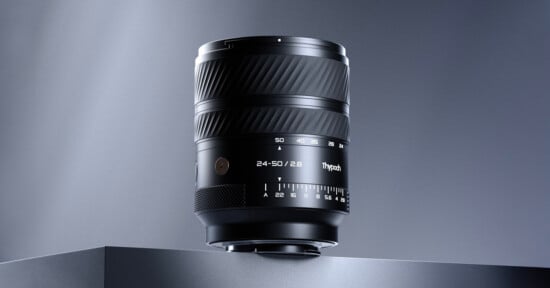 A black camera lens marked "24-50/2.8" and "TTArtisan" stands on a gray surface against a gradient gray background, with focus and aperture markings visible on its barrel.