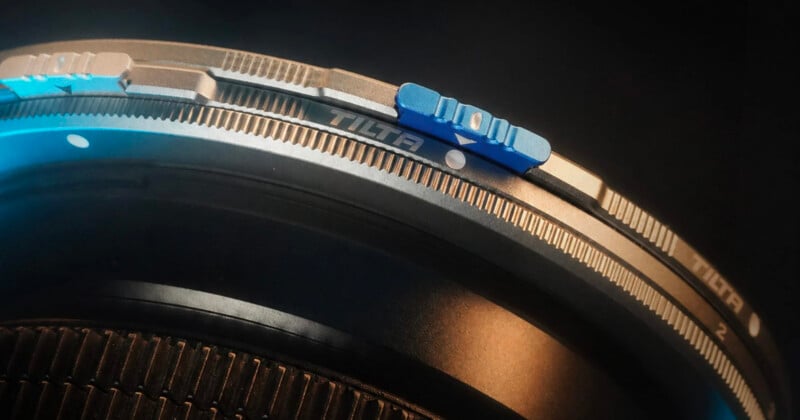 Close-up of a metallic camera lens accessory with engraved markings and a blue adjustment lever, featuring the brand name "Tilta" on the ring against a dark background.