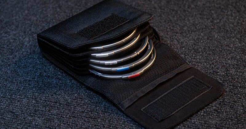 A black fabric case with several circular camera lens filters, each with colored markings, is open and displayed on a textured gray surface.