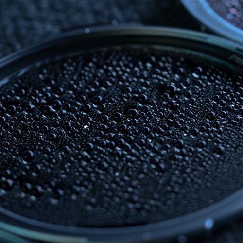 Close-up of a black surface covered with numerous small, shiny droplets, possibly condensation or liquid on a circular object, giving a textured and glossy appearance.