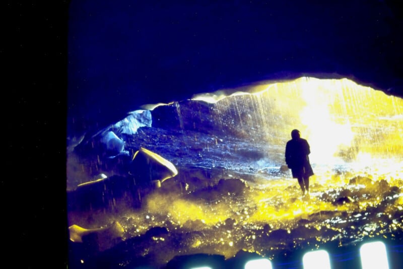 A silhouette of a person stands inside a cave, illuminated by bright yellow and blue light from the cave entrance, with rocky ground and mist creating a dramatic atmosphere.