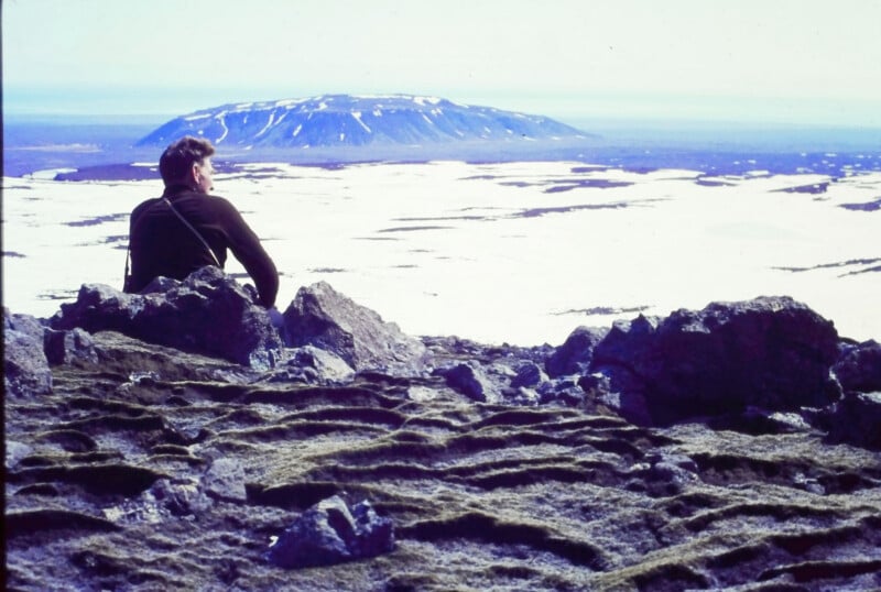 A person sits on rocky ground, overlooking a vast snowy plain with patches of snow and a distant, flat-topped mountain under a pale blue sky.