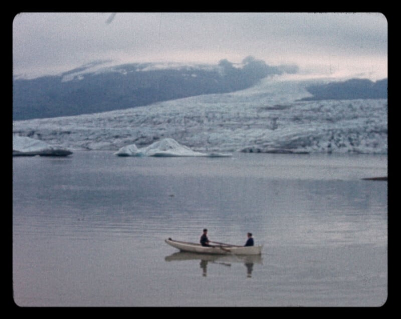 Two people row a small boat on a calm, icy lake with icebergs floating nearby. Snow-covered mountains and a glacier are visible in the misty background under a cloudy sky.