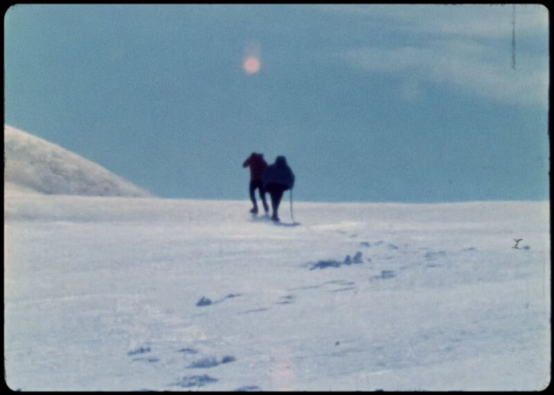 Two people in winter clothing hike up a snowy slope, leaving a trail of footprints behind them under a bright, clear sky.