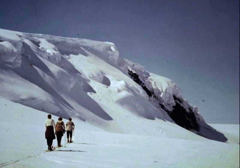 Four people walk in a line across a vast, snowy landscape toward a large, snow-covered mountain under a clear blue sky.