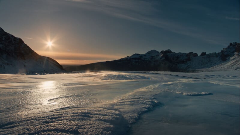 The sun sets over a frozen landscape, casting a golden glow on icy terrain with mountains in the background under a clear sky.