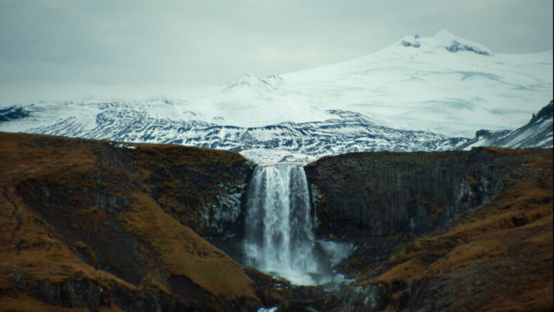 A tall waterfall cascades between rocky cliffs with brown grass, set against a backdrop of snow-covered mountains under a cloudy sky.
