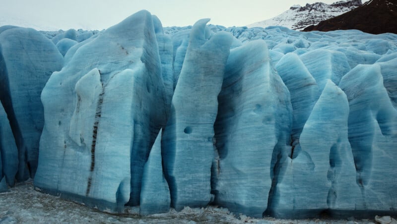Close-up view of tall, jagged blue glacier ice formations with textured surfaces, showing layers and cracks, set against a snowy mountain backdrop.