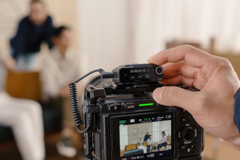 A close-up of a camera filming two people sitting and talking on a couch, with a hand adjusting an external microphone mounted on the camera. The camera screen displays the scene being recorded.