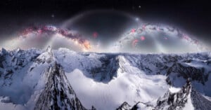 Snow-covered mountain peaks under a dark sky with bright, colorful arcs resembling galaxies and nebulae, creating a surreal and otherworldly landscape.