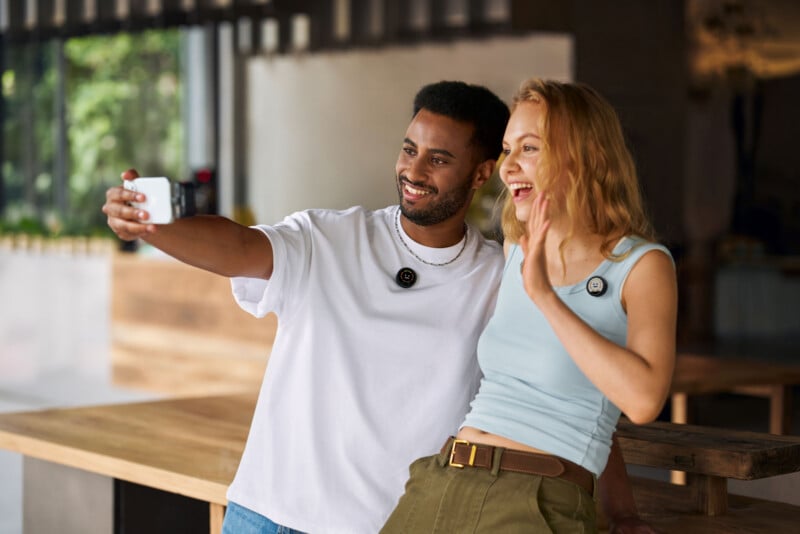 A man and a woman smile and pose for a selfie indoors. Both wear casual clothes and round electronic devices clipped to their tops. The woman waves, and they appear happy in a bright, modern setting.
