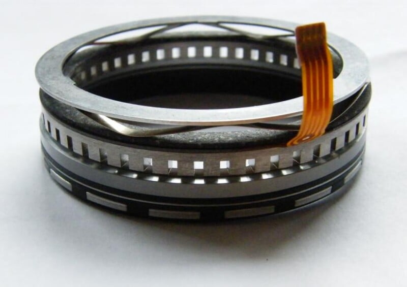 A close-up of an electronic rotary encoder with a metal ring, a flexible orange flat cable, and a segmented black and silver base, placed on a white surface.