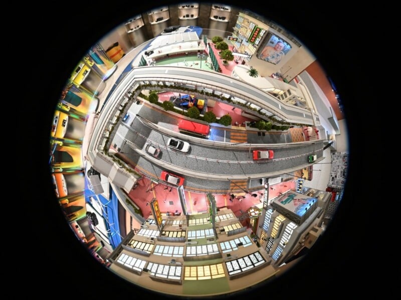 A fisheye lens view of a miniature city scene with roads, cars, buildings, and a pedestrian bridge, creating a circular, distorted perspective of an urban environment.
