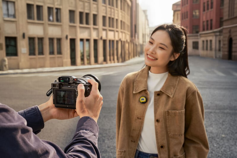 A person takes a photo of a smiling woman standing on an empty city street. She wears a tan jacket with a round yellow badge that says "Hi!" and poses with her hands in her pockets.