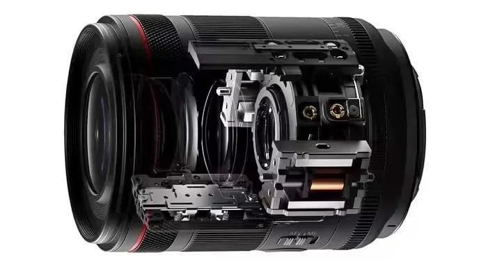 A camera lens with a partial cutaway view reveals its inner mechanical and optical components, showing intricate gears, glass elements, and electronic parts against a white background.