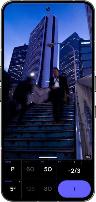 A smartphone screen shows a camera app capturing two blurred figures walking down urban stairs at dusk, with tall skyscrapers in the background and blue evening sky. Camera settings are visible at the bottom.