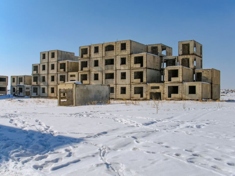 A large, unfinished concrete building stands abandoned in a snowy landscape under a clear blue sky, with footprints in the snow leading up to the structure.