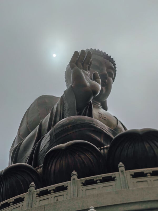 A large bronze Buddha statue with an upraised hand stands against a cloudy sky, with the sun faintly visible behind its head. The statue is surrounded by a decorative stone railing.