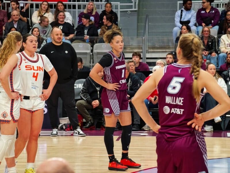 Basketball players stand on the court during a game, with one team in white and the other in maroon uniforms. Spectators fill the seats in the background, watching the action.