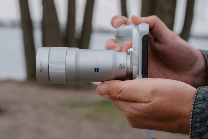 Close-up of a person holding a silver camera with a 400mm lens, outdoors near trees and water, with the camera pointed towards the right side of the image.