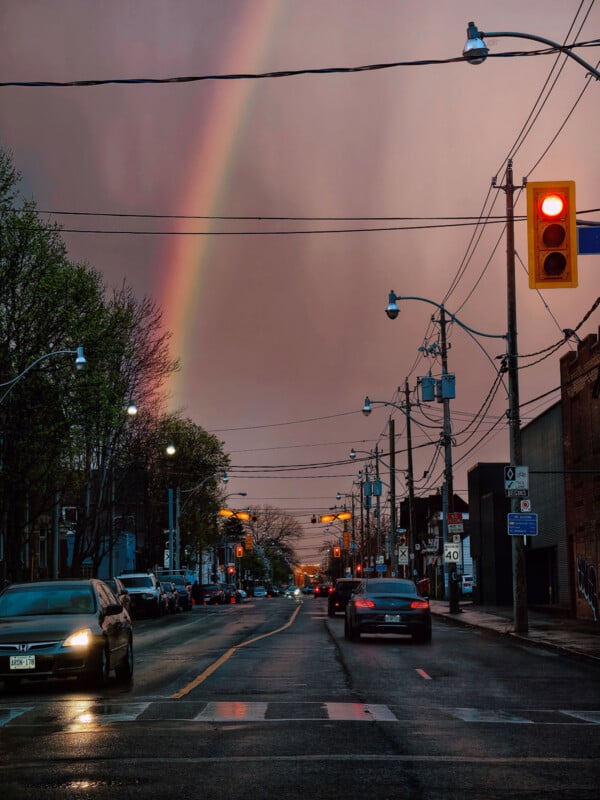 A city street at dusk with wet pavement, cars stopped at a red light, streetlights glowing, and a rainbow visible in the cloudy sky. Power lines and buildings line both sides of the road.