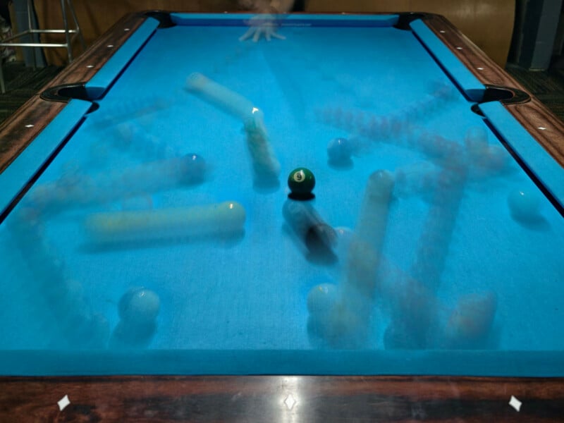 A pool table with multiple motion trails showing the movement of billiard balls and a player's hand, creating a dynamic, blurred effect against the blue felt surface.