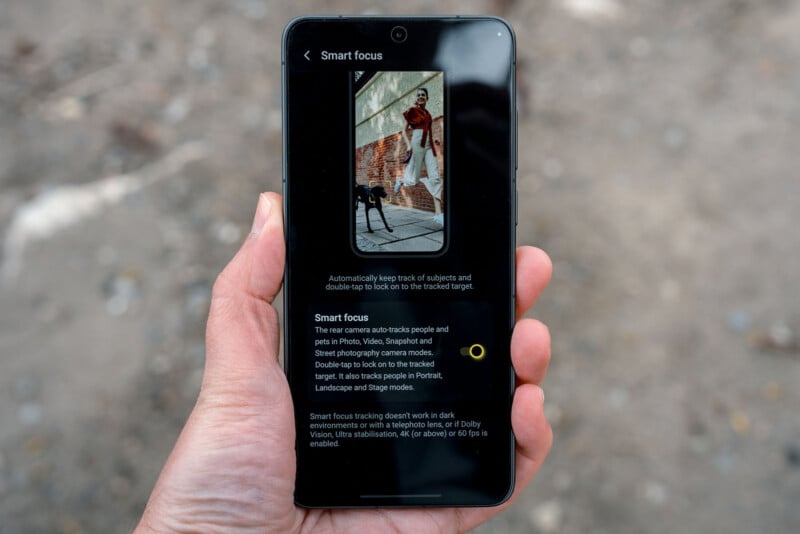 A hand holds a smartphone displaying a "Smart focus" camera feature, which explains how to auto-track subjects in photos and videos, with an image of a person and a dog on the screen. Outdoor ground is visible in the background.