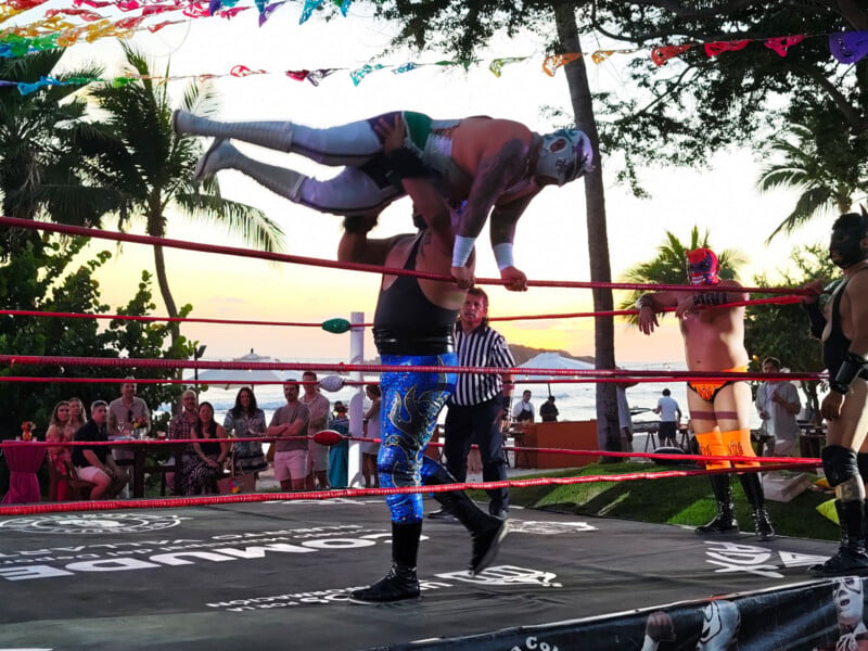 A masked wrestler in blue pants lifts another masked wrestler above his shoulders in a ring at sunset, while a referee and two other masked wrestlers look on, with palm trees and a crowd in the background.