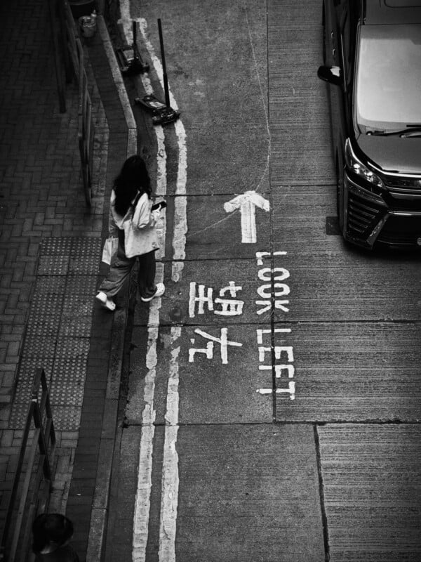 A woman walks along a sidewalk next to a street with markings that say "LOOK LEFT" and an arrow pointing left. Chinese characters are also painted on the road. A car is partially visible on the right. The image is in black and white.
