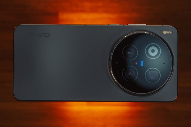 A black Vivo smartphone seen from the back, featuring a large circular triple camera module and flash, placed against a warm, glowing orange-brown background.