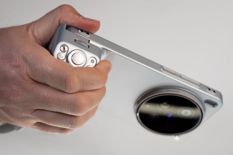 A hand holds a smartphone with a large round camera module and metallic grip attachment that resembles a camera, featuring buttons and dials for photography.