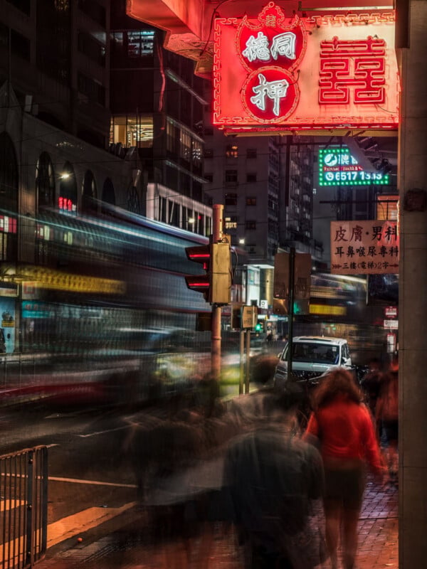 A busy city street at night with blurred motion of people walking, a passing double-decker bus, neon signs with Chinese characters, and illuminated buildings in the background.