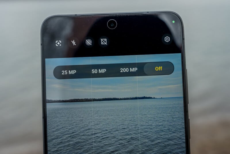 A smartphone camera screen displays photo resolution options: 25 MP, 50 MP, 200 MP, and Off. The background shows a large body of water with a distant shoreline under an overcast sky.