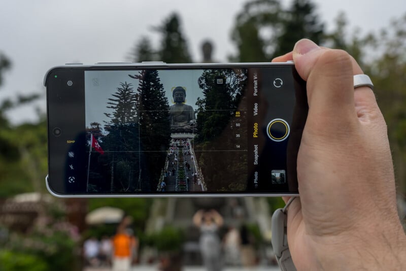 A hand holding a smartphone takes a photo of a large outdoor Buddha statue surrounded by trees and people, with the statue centered on the phone screen. The scene is slightly blurred in the background.