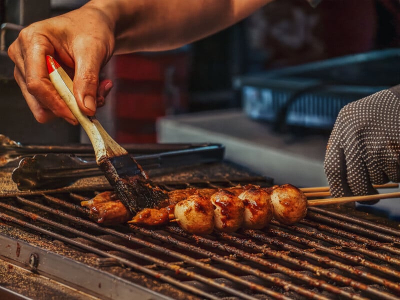 A person brushes sauce onto skewered grilled meatballs over a hot grill, with another hand holding tongs nearby. The scene appears to be at a street food stall.